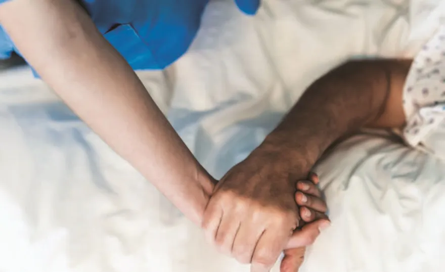 A cropped image of a nurse holding a patient's hand in a hospital bed. Only their linked hands and arms are visible.