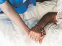 A cropped image of a nurse holding a patient's hand in a hospital bed. Only their linked hands and arms are visible.